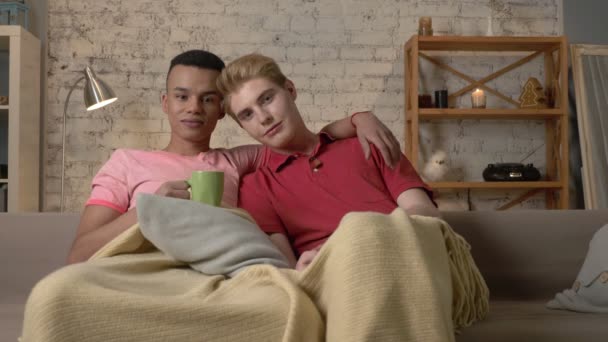 Twee homoseksuele jongens op de bank zitten en Tv kijken, drinken van hete thee, gebruik van de afstandsbediening. LGBT liefhebbers, een multinationale paar, een gelukkige homo familie, een thuis gezelligheid concept. Kijken naar de camera 60 fps — Stockvideo