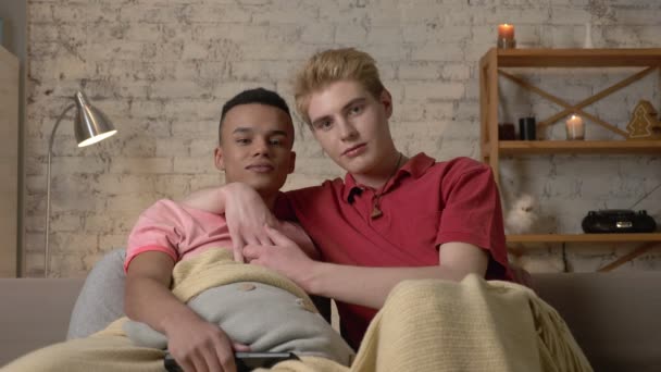 Два гея сидят на диване и смотрят телевизор, афроамериканец хочет переключить канал, но его останавливает американец. Любители ЛГБТ, многонациональная пара, счастливая семья геев, дом — стоковое видео