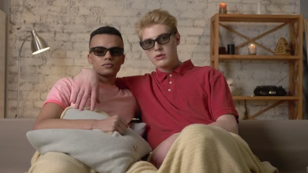 Two gay guys sit on a sofa covered in a warm blanket and watch a 3d scary movie, show an emotion of fright. LGBT lovers, happy gay family, home cosiness concept. Look at the camera 60 fps — Stock Video