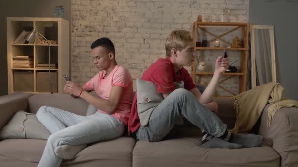 Two young men are sitting on the couch and using a smartphones, gays, the problem of society, a new generation, lgbt lovers, homo, homosexuality concept 60 fps — стоковое видео