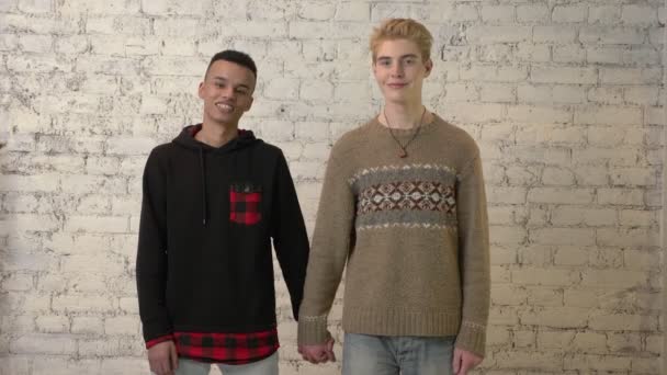 International gay couple holding hands, smiling, white brick wall on background. LGBT love, young couple, lover, homosexuality concept. 60 fps — Stock Video