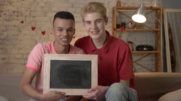 Una felice coppia gay internazionale è seduta sul divano e tiene un cartello vuoto. Comfort domestico sullo sfondo. 60 fps — Video Stock