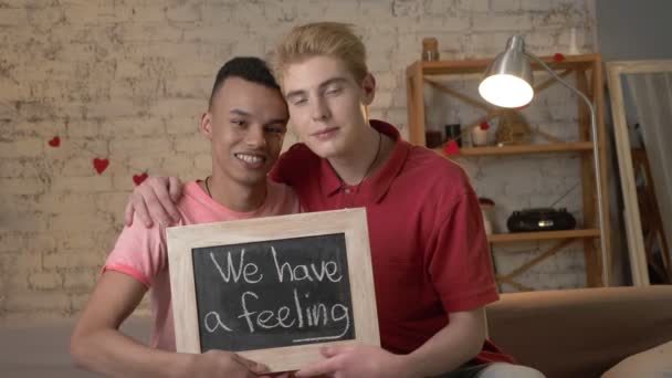 Szczęśliwa para gejów międzynarodowej jest siedzi na kanapie i posiadających znak. Mamy uczucie. Spójrz w stronę kamery. Domu na tle. 60 fps — Wideo stockowe