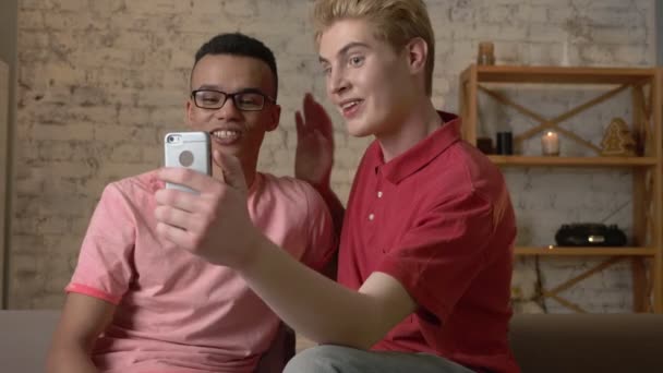 Dois amigos de diferentes nacionalidades usam um smartphone, mostram um gesto de saudação no chat por vídeo. 60 fps — Vídeo de Stock