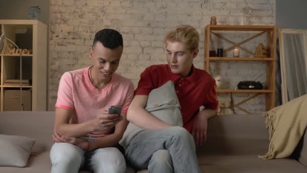 Dos amigos homosexuales internacionales se sientan en el sofá y ven fotos divertidas en el teléfono inteligente. Confort del hogar, familia, concepto de Internet. 60 fps — Vídeos de Stock