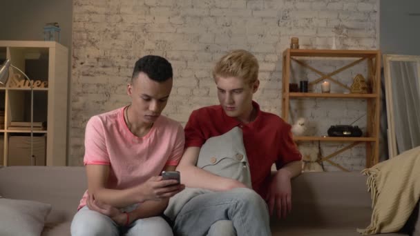 Dos amigos internacionales de un homosexual están sentados en el sofá y viendo fotos desagradables y mercenarias en un teléfono inteligente. Confort del hogar, familia, concepto de Internet. 60 fps — Vídeo de stock
