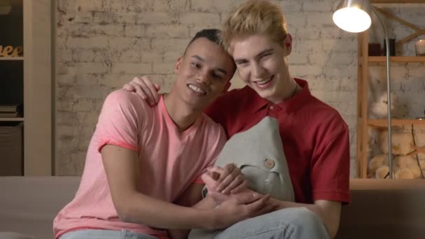 Two multi-ethnic homosexual friends sit on the couch holding hands, hugging, smiling, looking at the camera. Home cosiness, family, lgbt 60 fps — Stock Video