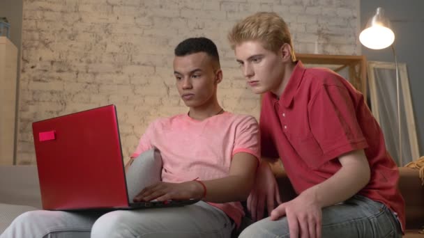 Twee multinationale homoseksuele vrienden op de bank zitten, een laptop gebruikt, kijken naar een interessante video op het Internet, verbaasd. De gezelligheid van het huis, familie, internet concept. 60 fps — Stockvideo