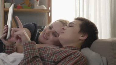 Lezbiyen çift kanepede dinleniyor, tablet bilgisayar kullanarak, tablet, gülümseyen, Aile İdil fotoğrafları kaydırma, sevgi, sevimli, 60 fps kadar yakın