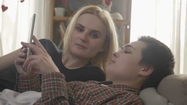 O casal de lésbicas está descansando no sofá, usando o computador tablet, rolando fotos no tablet, idílio familiar, amor, fofo, close. 60 fps — Vídeo de Stock
