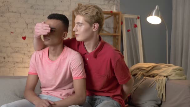 Un par de jóvenes homosexuales multinacionales se sientan en el sofá, un chico americano presenta un regalo a su pareja. Homeliness, gay, concepto de familia LGBT joven. 60 fps — Vídeos de Stock