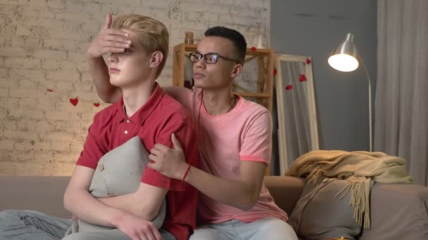 Пара молодых многонациональных гомосексуалистов сидит на диване, афроамериканец преподносит подарок своему партнеру. Гомосексуализм, геи, молодые ЛГБТ-семейные концепции. 60 кадров в секунду — стоковое видео