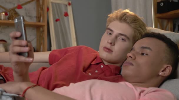 Um par de jovens homossexuais multinacionais deitou-se no sofá e usou um smartphone. Homeliness, gay, love, young LGBT family concept 60 fps — Vídeo de Stock