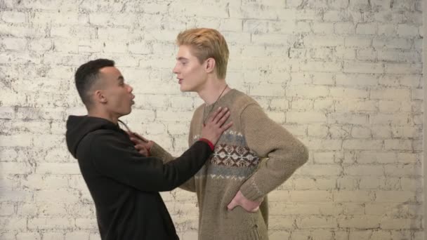 Молодой американец со светлыми волосами пытается поцеловать своего африканского друга, но он против. ЛГБТ, гомосексуализм, гомосексуальность. 60 кадров в секунду — стоковое видео