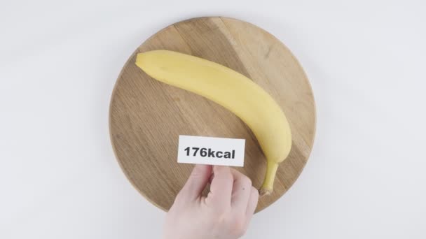 Quantità di calorie in banana, mano maschile mette un piatto con il numero di calorie su una banana, colpo superiore 60 fps — Video Stock