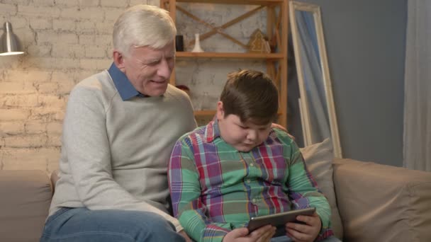 Dědeček a vnuk sedí na gauči, používání tabletu a při pohledu na fotoaparát. Pohodlí domova, rodiny idyly, útulnost koncept. 60 snímků za sekundu — Stock video
