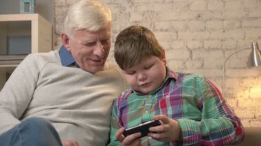 Dedesi ve bir smartphone kullanarak kanepede oturan torunu bahsetmişken, şişman bir çocuk Smartphone'da oynuyor. Dede torunu için sevinir. Ev konfor, rahatlık iletişim, Aile İdil