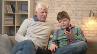 Dede ve torunu bir smartphone kullanarak kanepede oturuyor, selfie yap. Genç şişman çocuk ve büyükbaba. çocuk barış işareti gösterir. Ev konfor, Aile İdil, rahatlık kavramı. 60 fps