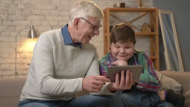 Grand-père et petit-fils sont assis sur le canapé à l'aide d'une tablette, agitant la main dans le chat vidéo, saluant, souriant et riant. Confort à la maison, idylle familiale, confort, internet, concept vidéo. 60 ans — Video