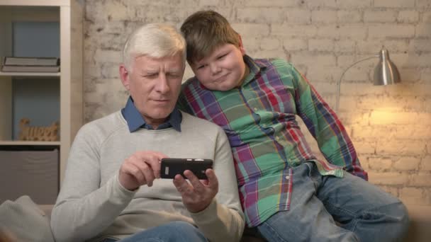 Grand-père et petit-fils sont assis sur le canapé à l'aide d'un smartphone, jouer sur un smartphone. Jeune gros garçon et grand-père. Confort à domicile, idylle familiale, concept de confort. 60 ips — Video