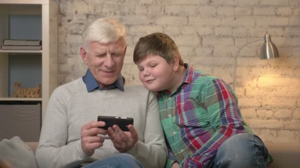 Grandfather and grandson are sitting on the couch using a smartphone, playing on a smartphone. Young fat boy and grandfather. Home comfort, family idyll, cosiness concept 60 fps — Stock Video