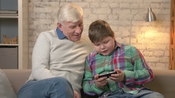 Дедушка и внук сидят на диване, используя смартфон, толстый ребенок играет на смартфоне. Молодой толстяк и дедушка. Домашний комфорт, семейный уют, понятие уюта. 60 кадров в секунду — стоковое видео