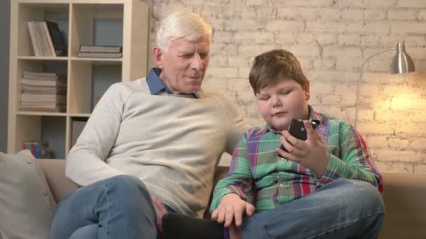 Grandfather and grandson are sitting on the sofa using a smartphone, do selfie. Young fat child and grandfather. boy shows the sign of peace. Home comfort, family idyll, cosiness concept. 60 fps — Stock Video
