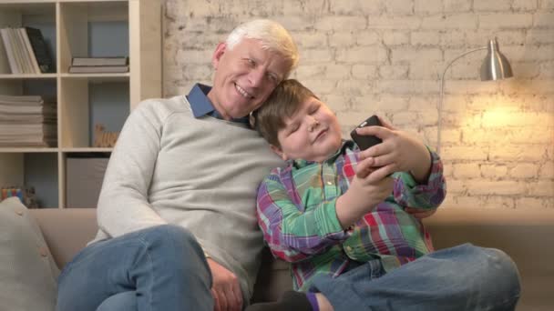 Großvater und Enkel sitzen mit dem Smartphone auf dem Sofa, machen Selfie. kleines dickes Kind und Großvater lächelnd, schmusend. Wohnkomfort, Familienidylle, Gemütlichkeit. 60 fps