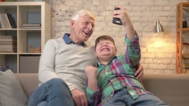 Grand-père et petit-fils sont assis sur le canapé à l'aide d'un smartphone, faire du selfie. Réjouissez-vous de la victoire, riez, ils sont heureux. Jeune gros enfant et grand-père. 60 ips — Video