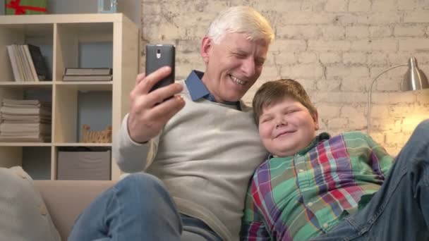 Grand-père et petit-fils sont assis sur le canapé. Le vieil homme tient un smartphone dans sa main, faisant un selfie. Ils rient, ils sont heureux. Jeune gros enfant et grand-père. 60 ips — Video