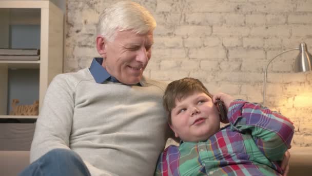 Grand-père et petit-fils sont assis sur le canapé. Un jeune gros garçon parle sur un smartphone. Confort à la maison, dialogue, idylle familiale, concept de confort. 60 ips — Video