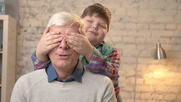 Fiatal kövér gyerek becsukja a nagyapák szemét a kezével. Találd ki. Egy játék, egy tréfa. Boldog családi koncepció. Otthoni kényelmet, családi idill nyugalma koncepció. 60 fps — Stock videók