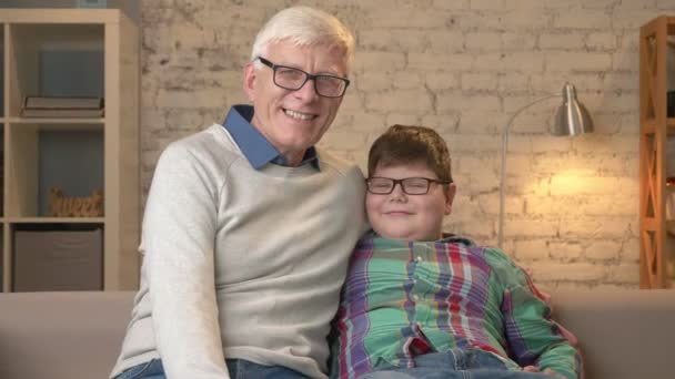 Abuelo y nieto con gafas, sentado en el sofá, sonriendo y mirando a la cámara. Confort en el hogar, idilio familiar, concepto de comodidad 60 fps — Vídeos de Stock