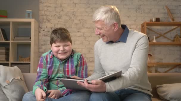 Пожилой человек учит молодого толстяка. Дедушка и внук читают интересную книгу, улыбаются, счастливая семья, концепция домашнего уюта. 60 кадров в секунду — стоковое видео