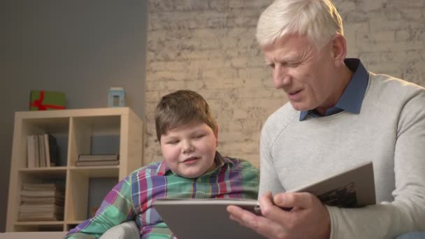 Oudere man leert een jonge dikke kerel om te lezen. Opa en kleinzoon, gelukkige familie, thuis gezelligheid, opleiding, kennis, lessen concept. 60 fps — Stockvideo