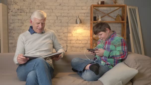 A diferença de gerações. Homem idoso está sentado no sofá e lendo um livro, um jovem gordo usa um tablet e fones de ouvido, falando. Conforto em casa, idílio familiar, conceito de conforto 60 fps — Vídeo de Stock