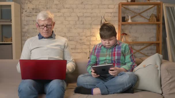 Het verschil tussen de generaties. Oudere man met bril zittend op de Bank bezig met laptop, vet jongen speelt op tablet. Home comfort, familie idylle, gezelligheid concept 60 fps — Stockvideo