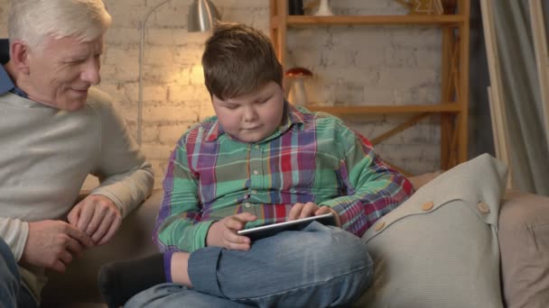 Young fat boy is sitting with his grandfather on the couch and playing on the tablet. The difference of generations. Home comfort, family idyll, cosiness concept. Clode up 60 fps — Stock Video