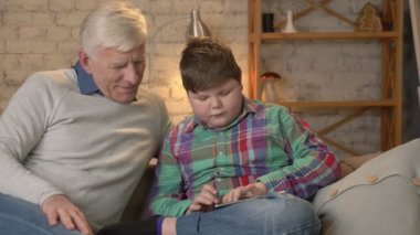 Genç şişman çocuk onun dedesi ile kanepede oturan ve tablet üzerinde oynama. Nesil farkı. Ev konfor, Aile İdil, rahatlık kavramı. 60 fps