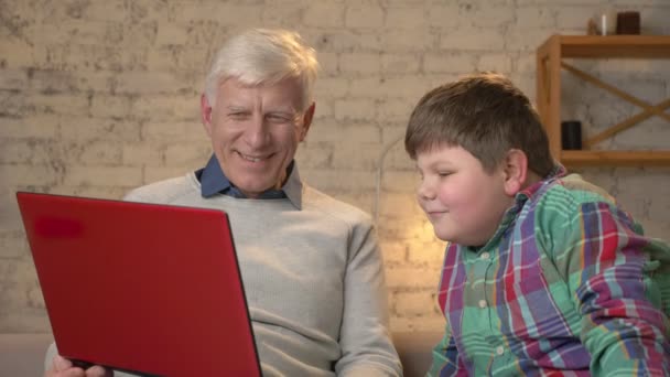 Παππούς και εγγονός κάθονται στον καναπέ και παρακολουθούν μια αστεία ταινία για το φορητό υπολογιστή. Αρχική idyll οικογένειας, άνεση, ζεστασιά έννοια, η διαφορά των γενεών 60 fps — Αρχείο Βίντεο