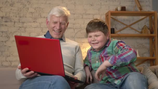 Grandfather and grandson are sitting on the couch and watching a funny movie on the laptop, laughing. Home comfort, family idyll, cosiness concept, difference of generations 60 fps — Stock Video