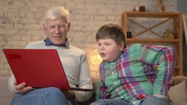 Дедушка и внук сидят на диване и смотрят забавный, интересный фильм на ноутбуке, смеясь. Домашний комфорт, семейный уют, концепция уюта, смена поколений — стоковое видео