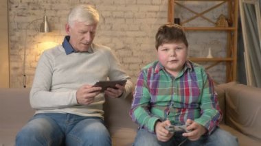 Dede ve torunu kanepede oturuyor. Yaşlı bir adam anlamları bir tablet, genç bir şişman adam konsol oyun oynar, kaybeder. Video oyunları. Ev konfor, Aile İdil, rahatlık kavramı, farkı
