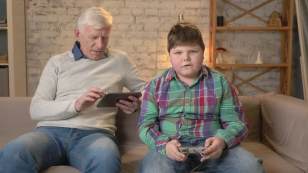 Nagyapa és unokája ül a kanapén. Egy öreg ember használ egy tabletta, egy fiatal kövér srác játszik a konzol játék. Videojátékok. Otthoni kényelmet, családi idill, nyugalma koncepció, különbség — Stock videók