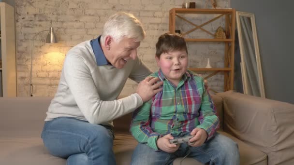 El abuelo disfruta de la victoria de su nieto en el juego de consola. El viejo está sentado en el sofá con un chico gordo. Videojuego. confort en el hogar, idilio familiar, concepto de comodidad, diferencia de — Vídeo de stock