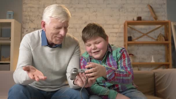 El nieto enseña al abuelo a jugar el juego de consola. El anciano está aprendiendo a jugar un videojuego, no lo entiende. confort en el hogar, idilio familiar, concepto de comodidad, diferencia de — Vídeo de stock