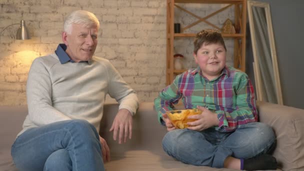 Дедушка и его внук сидят на диване и смотрят телевизор, толстый мальчик предлагает дедушке поесть чипсов, он не согласен. Домашний комфорт, семейный уют, понятие уюта, разность — стоковое видео