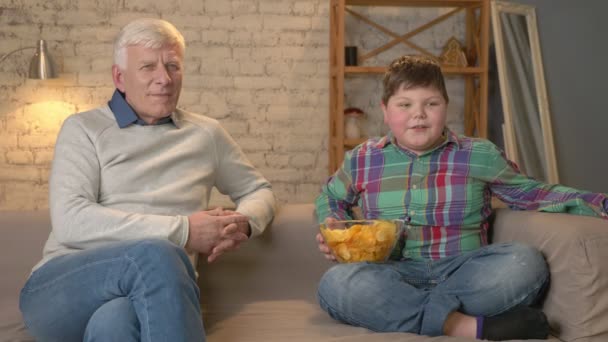 Дедушка и его внук сидят на диване и смотрят телевизор, едят чипсы, улыбаются. Пожилой человек переключает каналы, использует пульт дистанционного управления. Домашний комфорт, семейный уют, уютность — стоковое видео