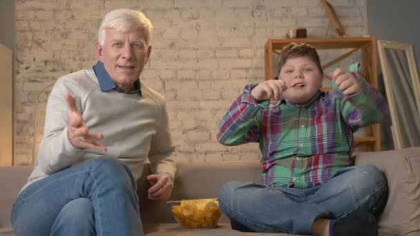 Dědeček a vnuk se sedí na gauči a dívat na televizi, jíst chipsy, těší vítězství, diváky, s úsměvem. Pohodlí domova, rodiny idyly, útulnost koncepce, rozdíl generací. 60 snímků za sekundu — Stock video