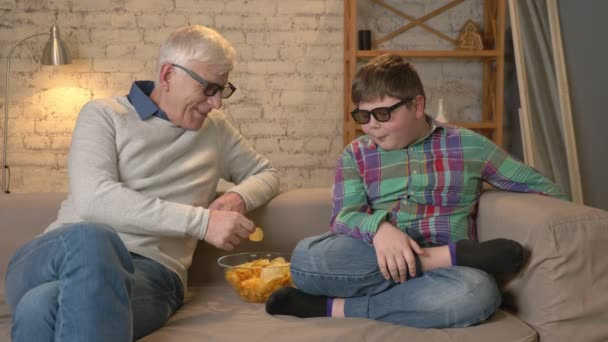Grandfather is feeding his grandson with chips from his hands. An elderly man and a young fat boy are sitting on the couch in 3d glasses. Home comfort, family idyll, cosiness concept, difference of — Stock Video